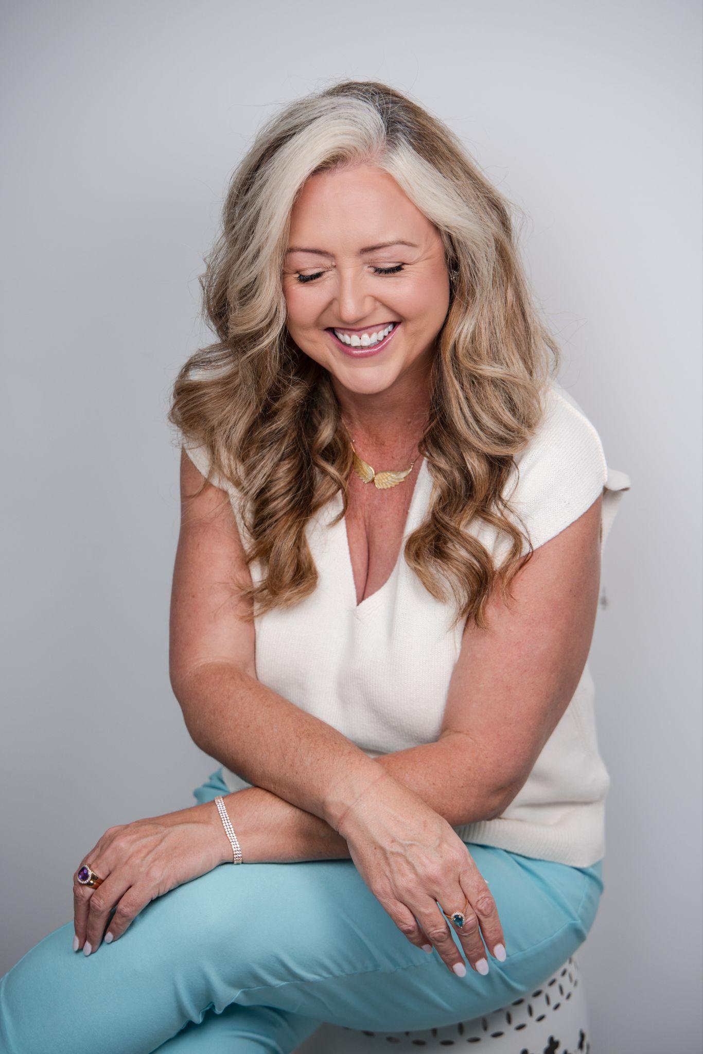 Smiling woman with long, wavy hair sitting on a stool, wearing a light top