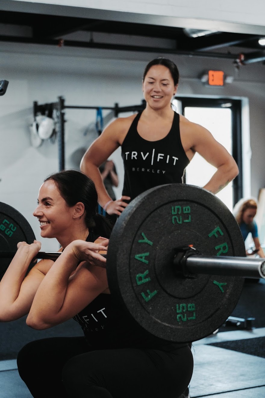 Two women in fitness clothing, one lifting weights, the other providing encouragement in a gym