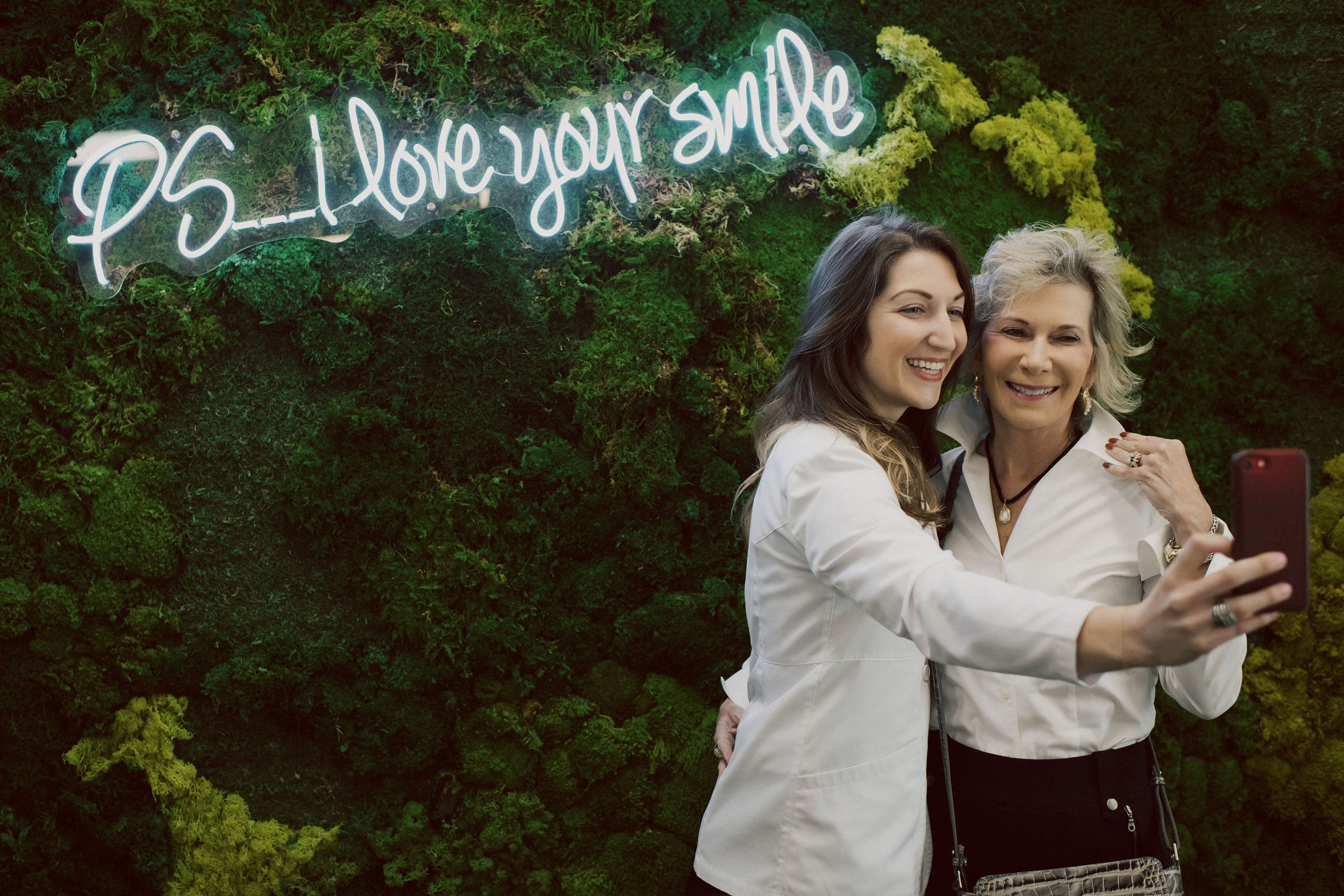 Two women smiling and taking a selfie in front of a green moss backdrop.