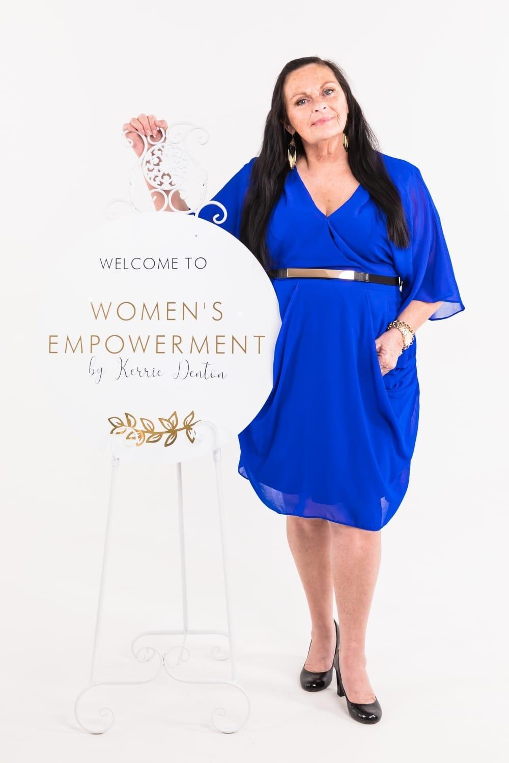 A woman in a blue dress stands beside a sign for a women's empowerment event