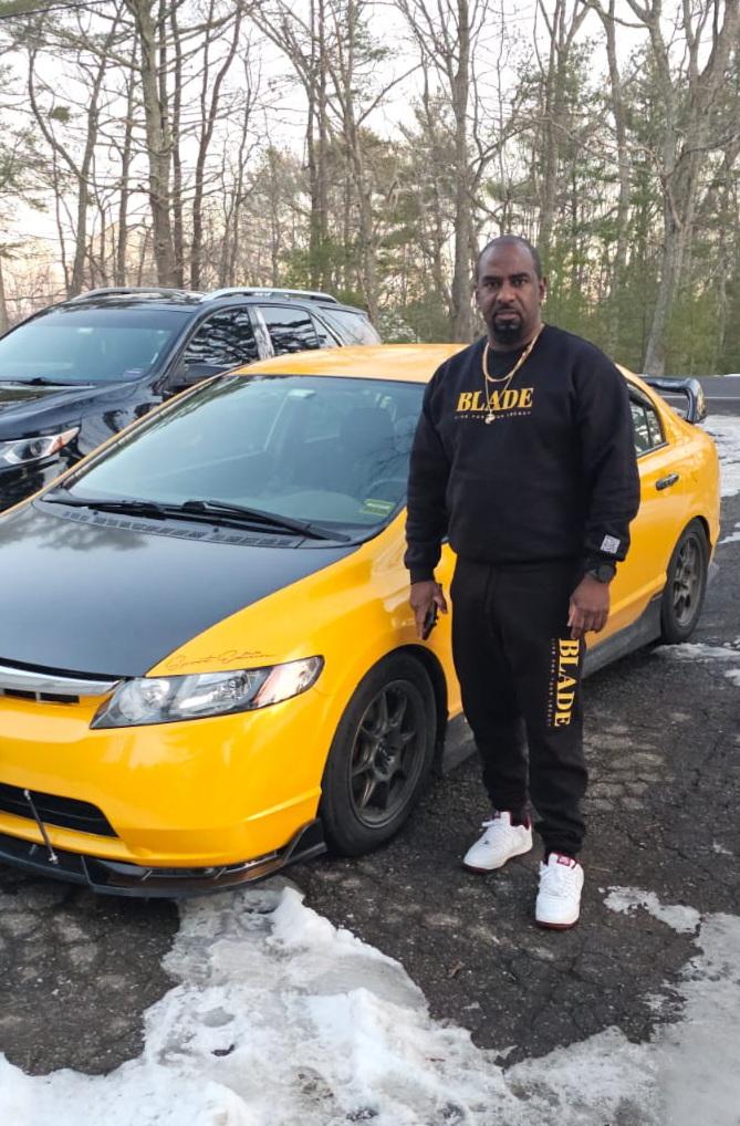 Man stands beside a yellow car wearing a matching black outfit with "BLADE" logo