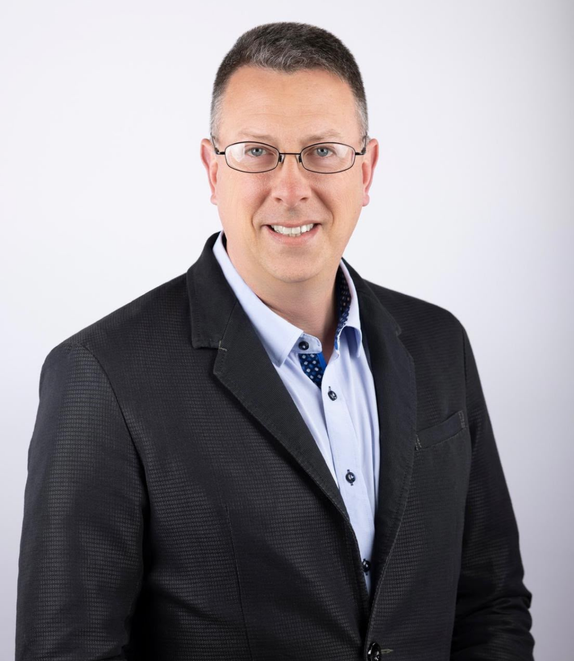 Smiling man in a black blazer and light blue shirt against a plain background
