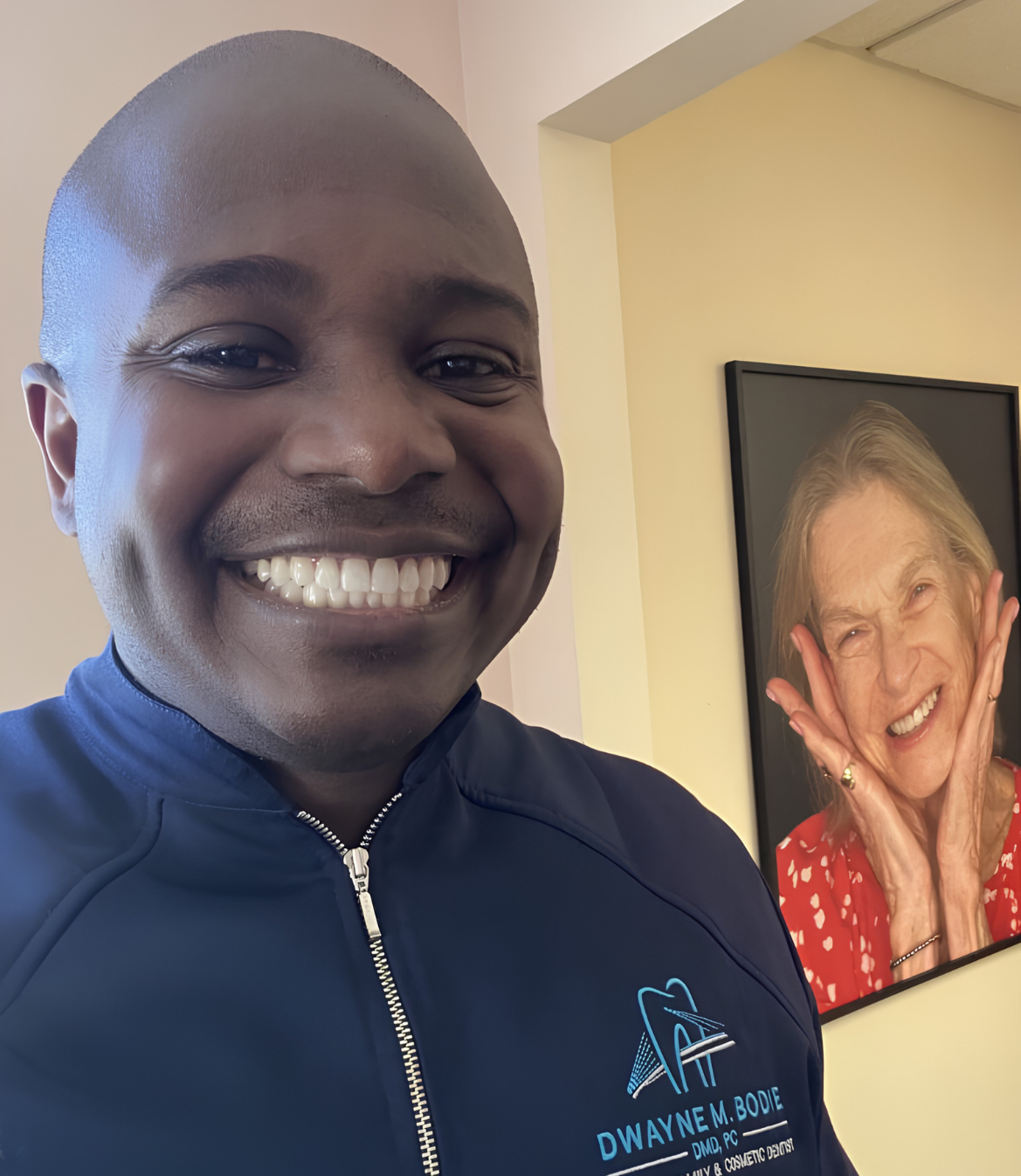 Smiling man in a dental coat poses near a portrait of a joyful woman