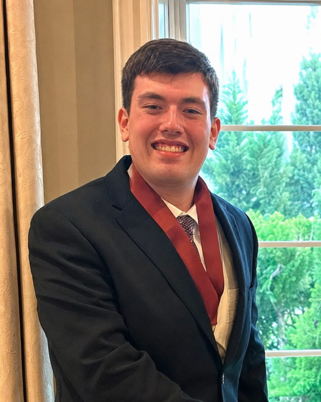 A young man in a suit smiles, wearing a maroon sash, near a window.