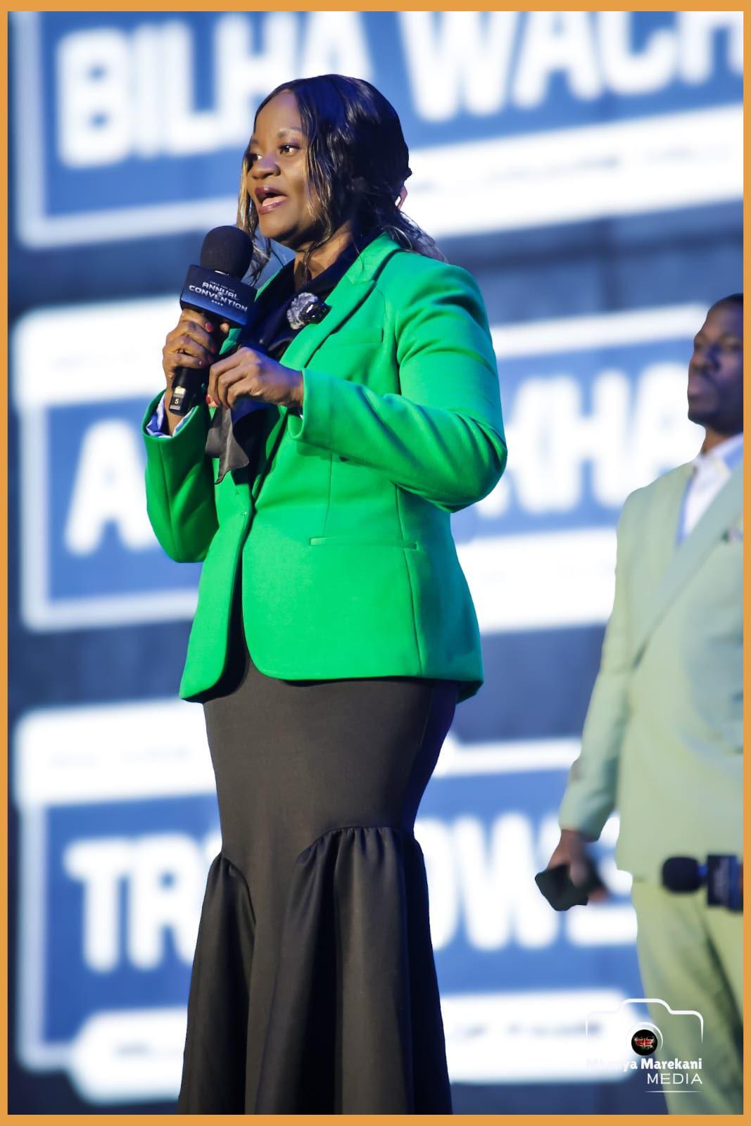Woman in a green blazer speaks into a microphone on stage at an event
