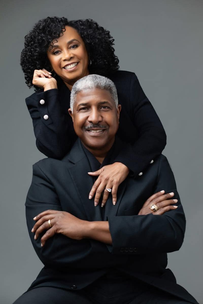 A smiling woman with curly hair poses behind a man in a black suit.