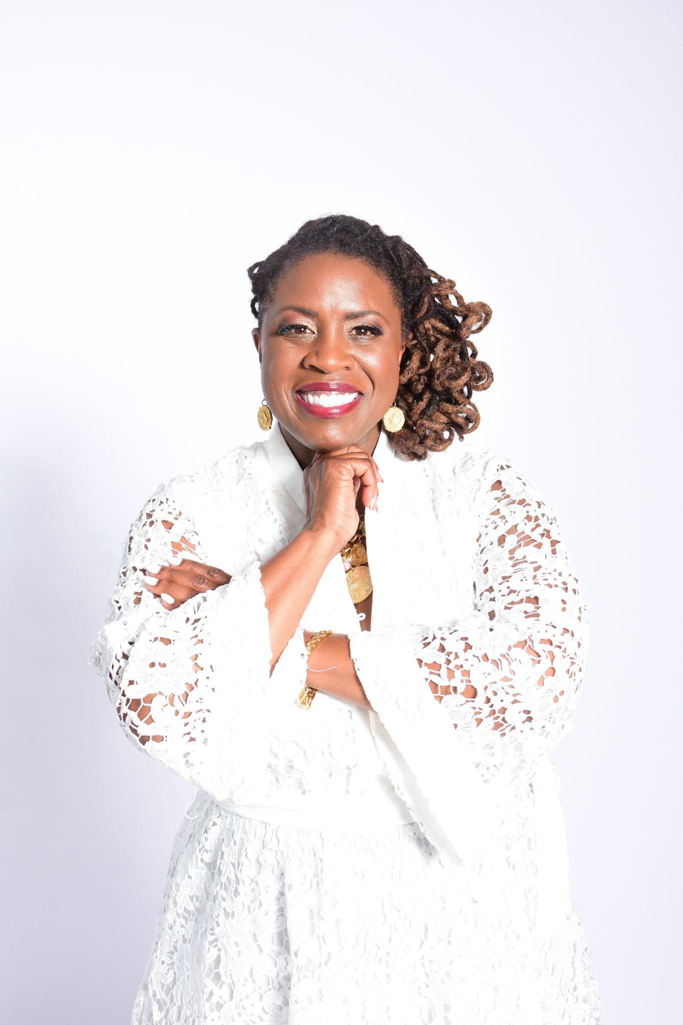 A woman in a white lace dress poses confidently with arms crossed and a smile