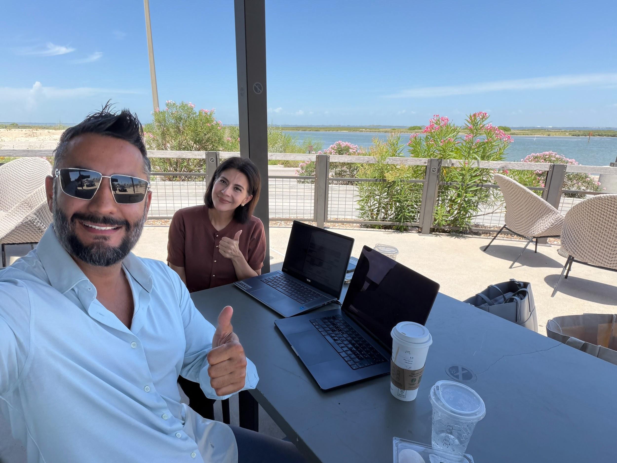 Two people smiling at a table with laptops and drinks, outdoors by a water view