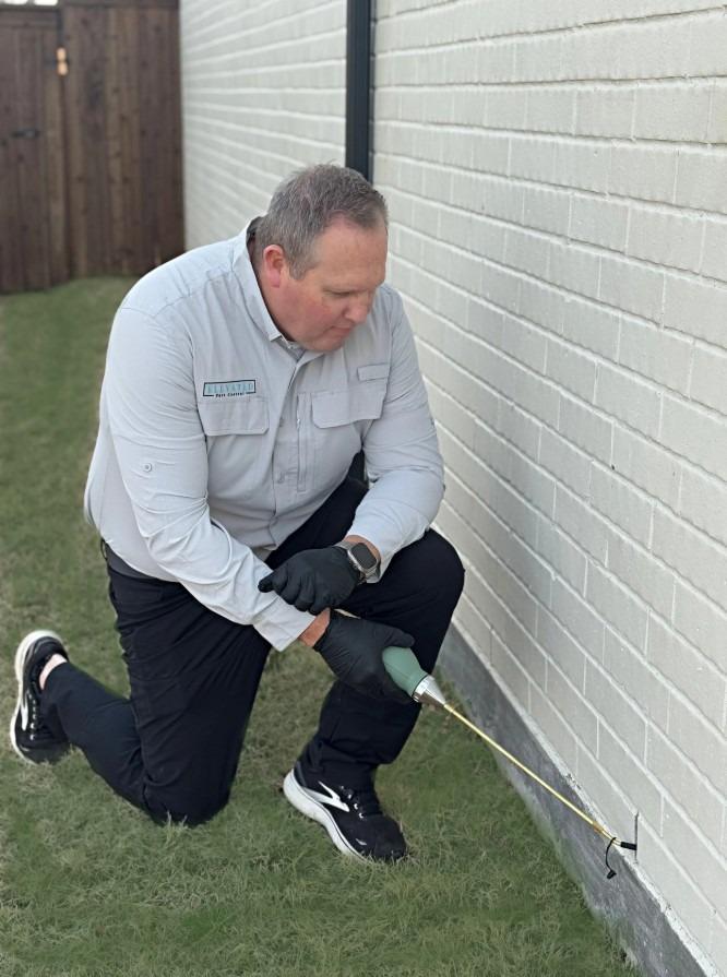 Person kneeling beside a white wall, using a spray tool for pest control