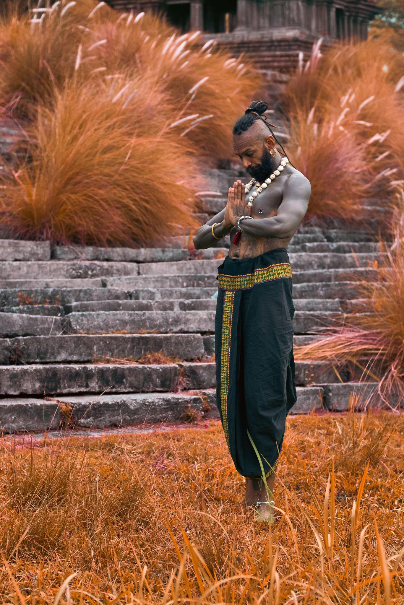 Person with a prayer pose in traditional attire amidst tall grass and stone steps