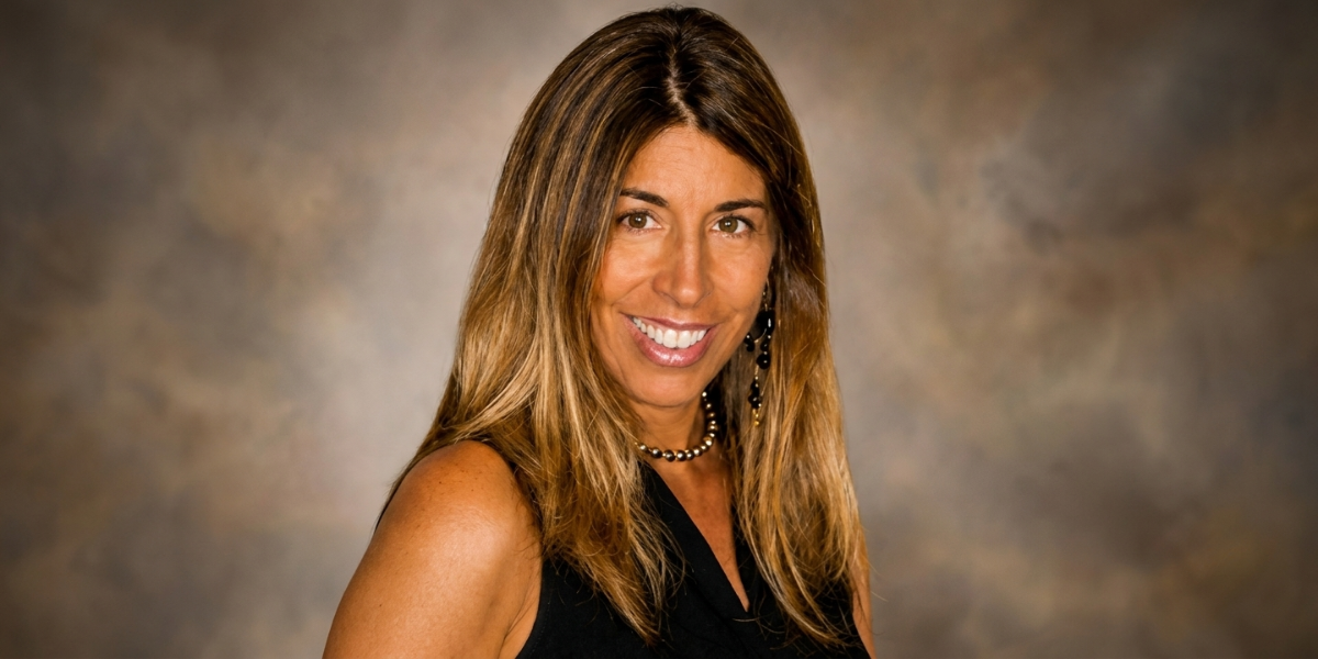 Woman with long, wavy brown hair smiling in a black outfit against a blurred background