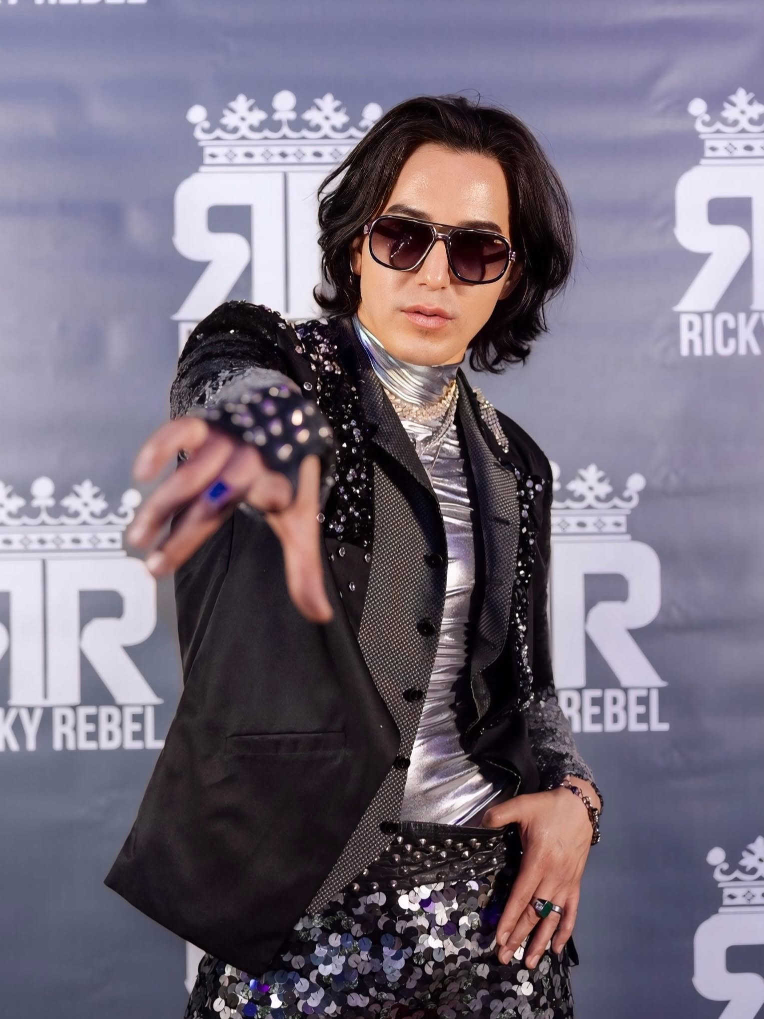 Person in sunglasses and sequined outfit posing confidently against a branded backdrop