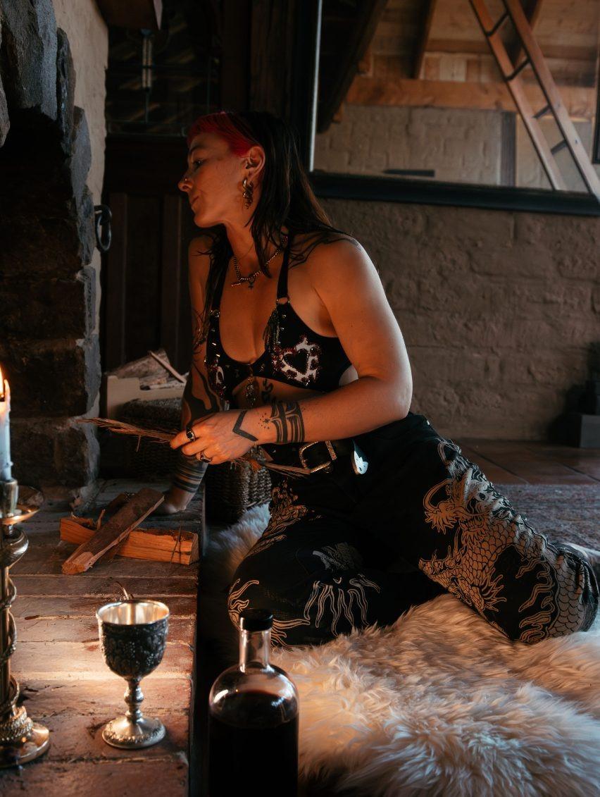 Woman in a dark, stylish outfit kneels by a stone fireplace, surrounded by candles and decor.