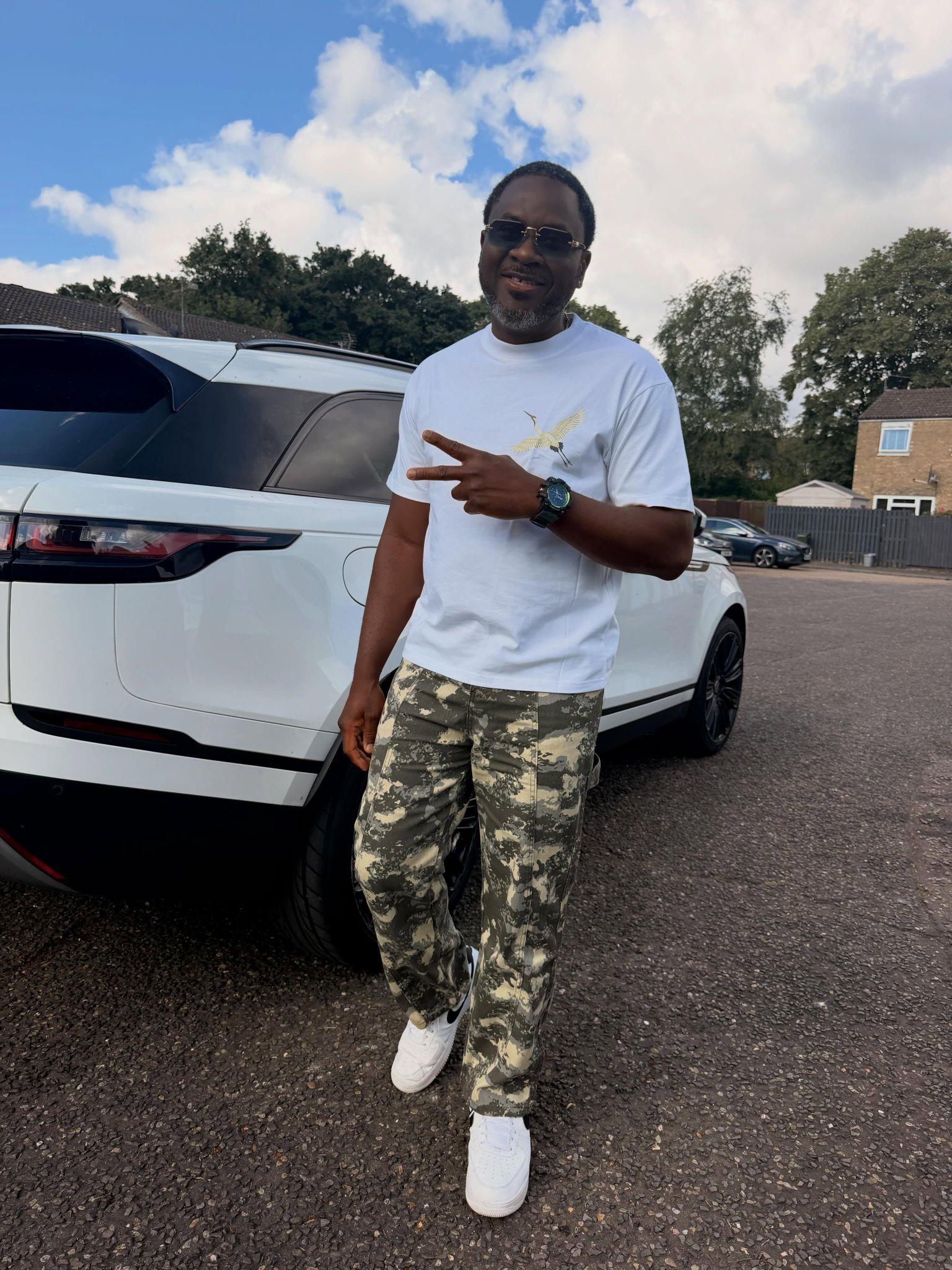 Man in a white t-shirt and camouflage pants stands by a white SUV