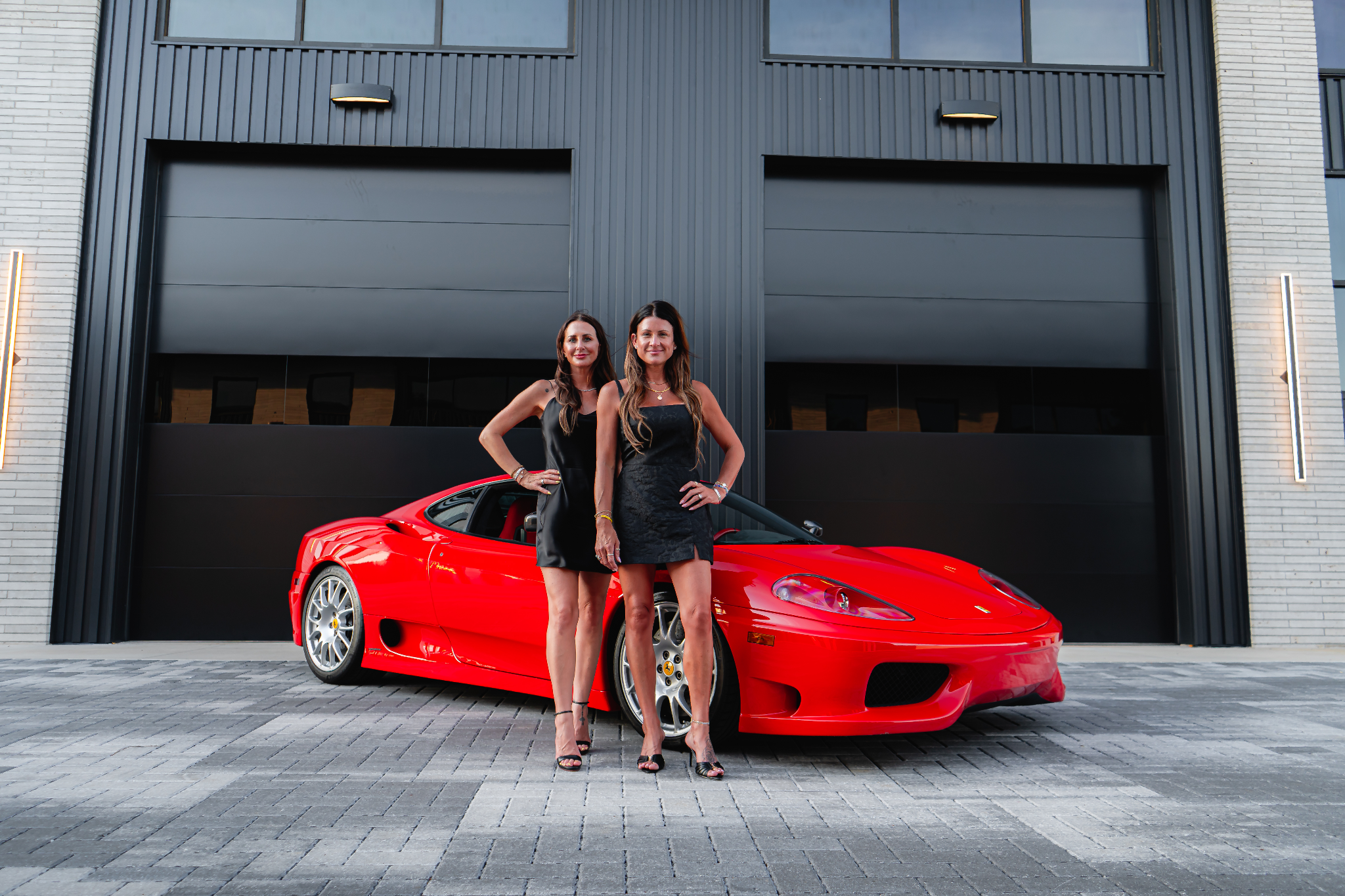 Two women in black dresses standing next to a red sports car outdoors