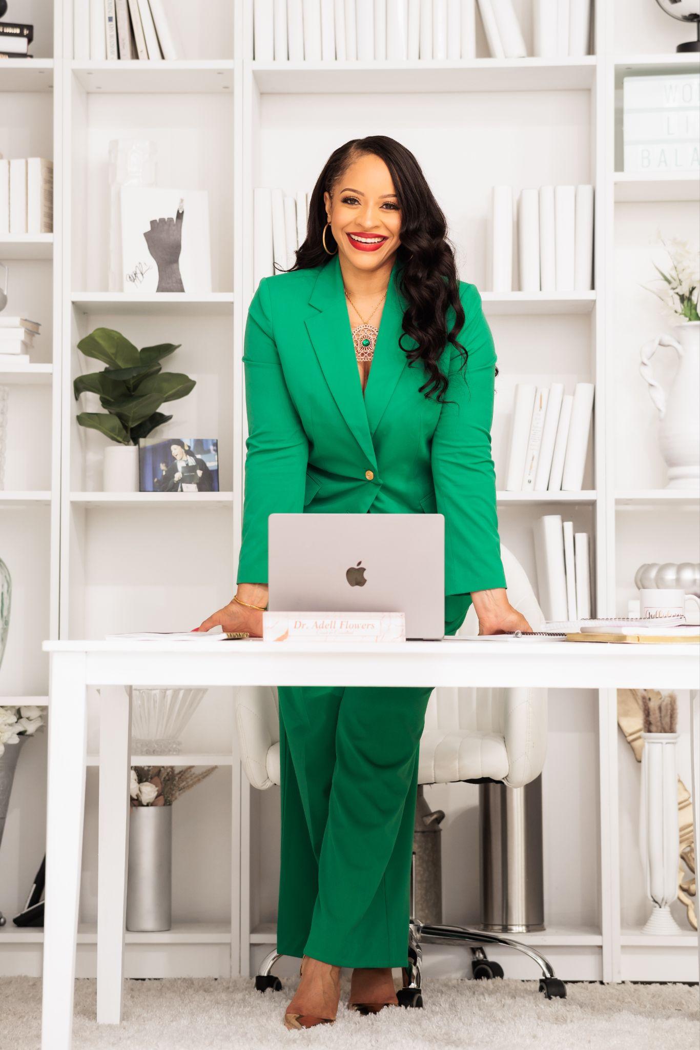 Woman in a green suit standing by a desk with a laptop and bookshelf backdrop