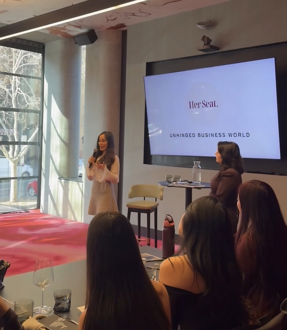 A woman speaks at an event titled "Her Seat: Unhinged Business World" while others listen.