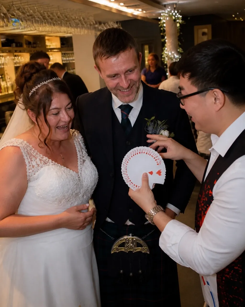A magician performs a card trick for a smiling bride and groom at a wedding.
