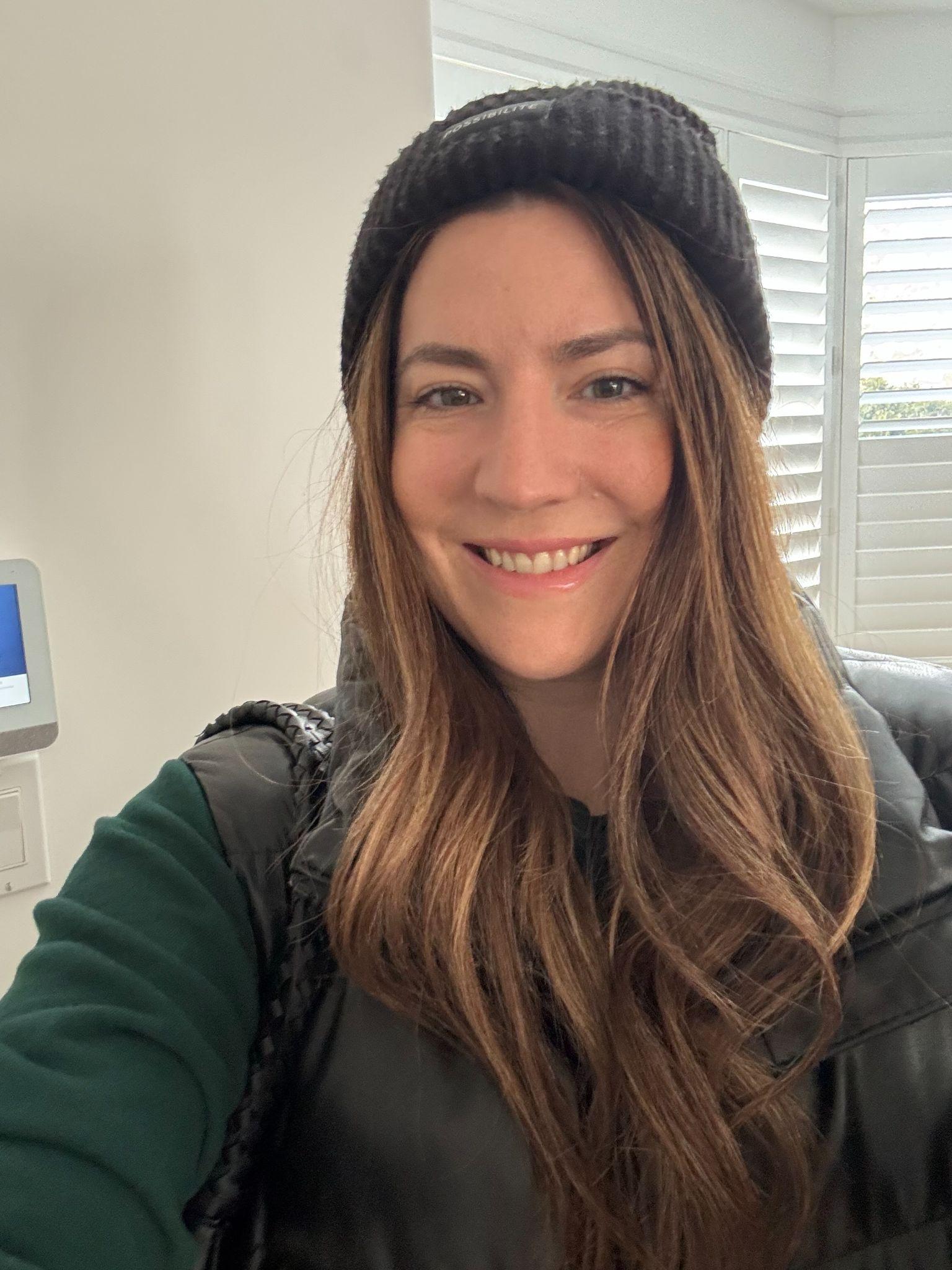 A woman with long hair and a beanie smiles in a bright room