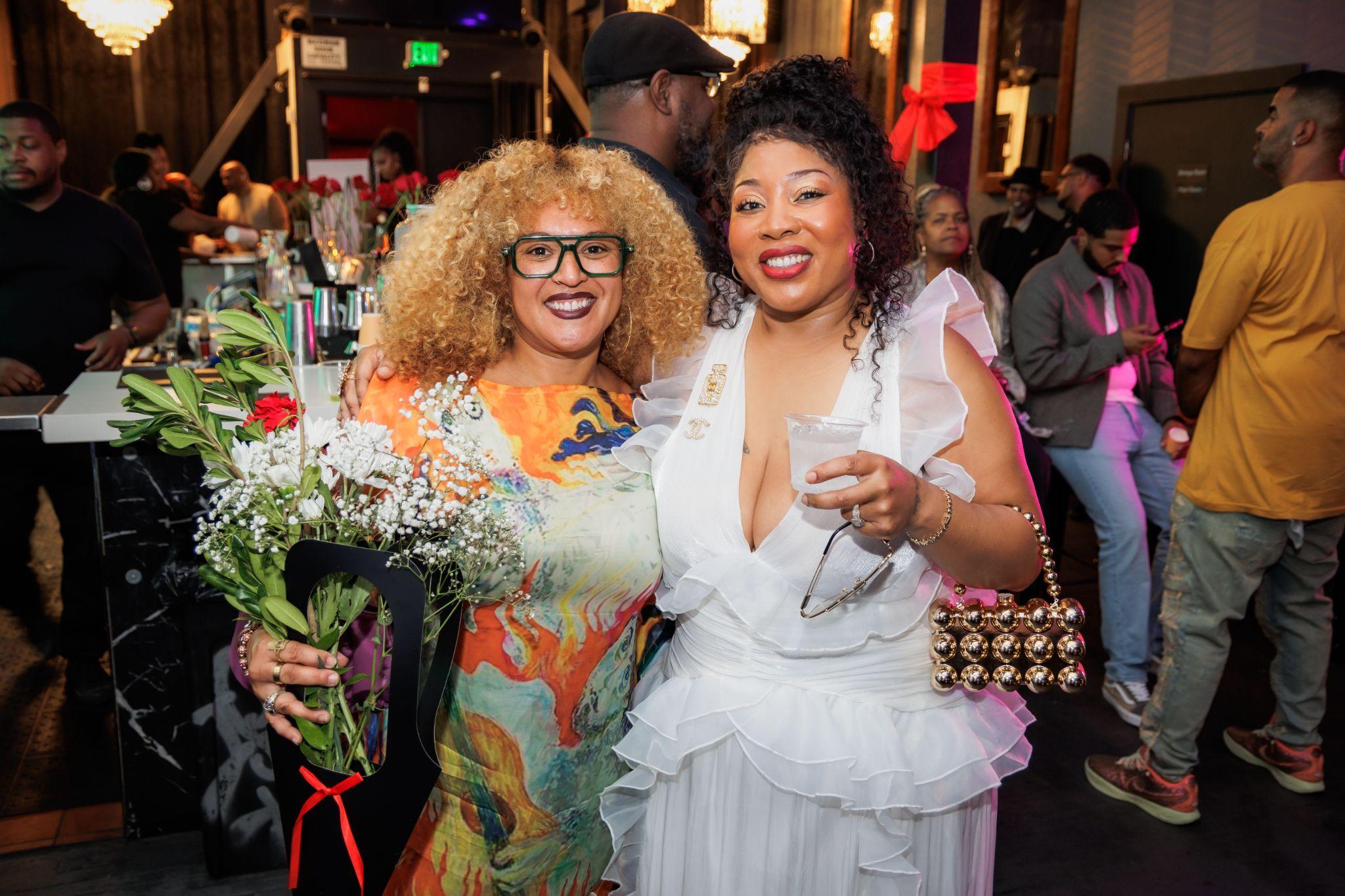 Two women smiling together at a lively event, one holding flowers and a drink