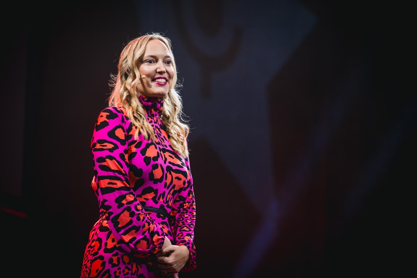 A woman with long hair in a bold pink and black dress smiles on stage.