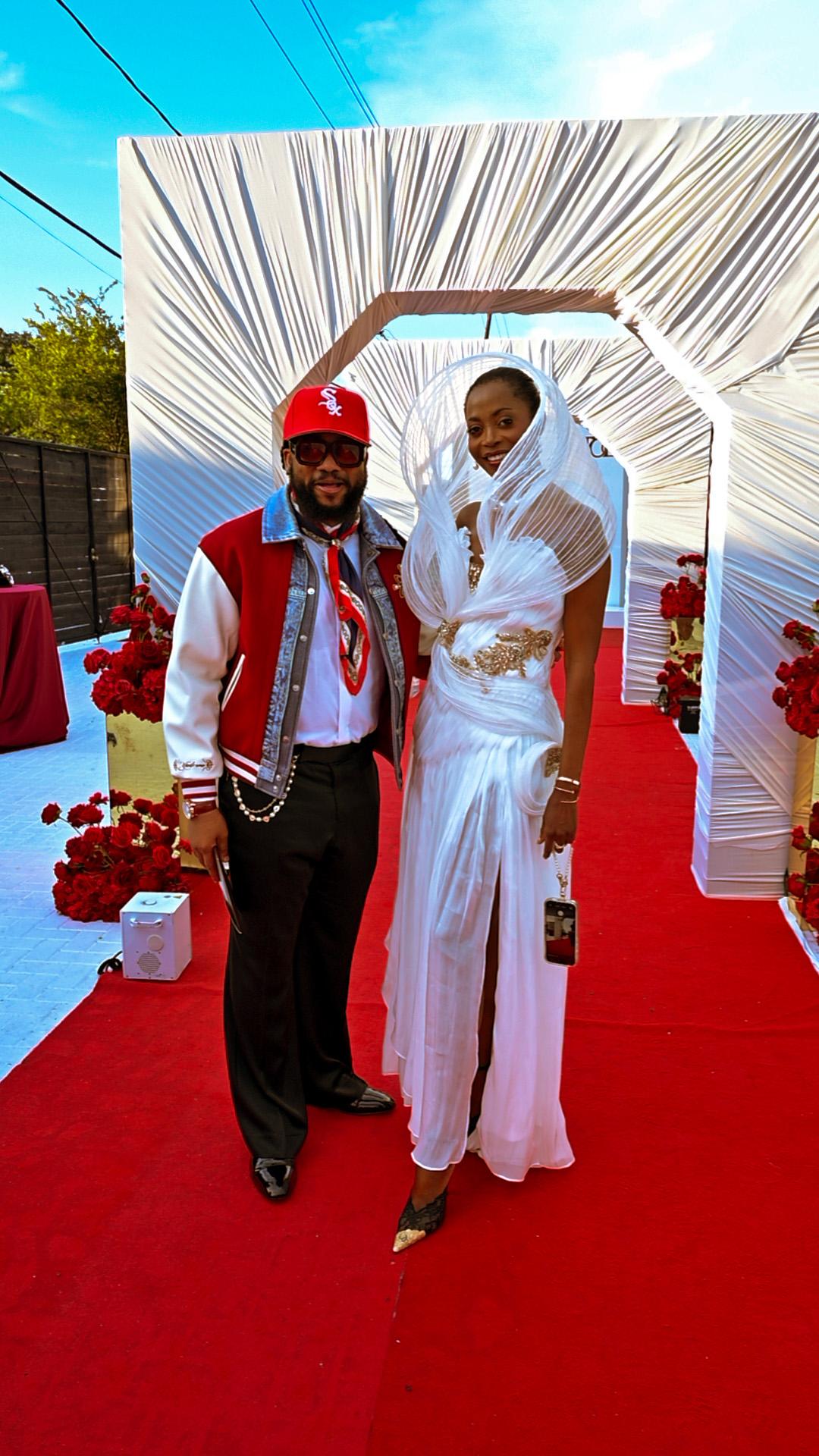 Couple posing on a red carpet, dressed in elegant outfits with floral decorations nearby
