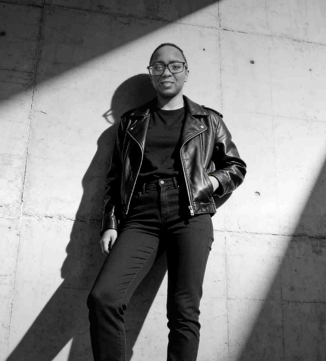 Young woman in a leather jacket stands confidently against a concrete wall