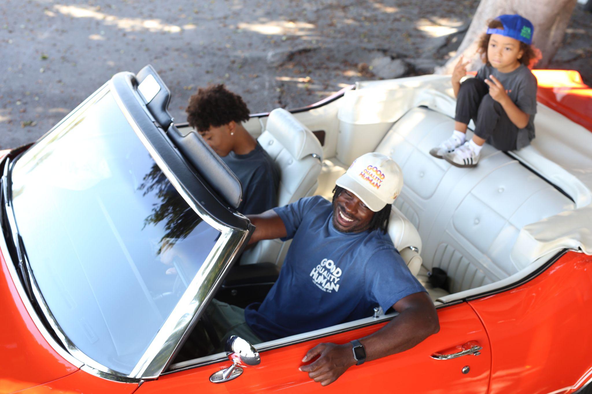 A man in a convertible smiles, accompanied by a woman and a child.