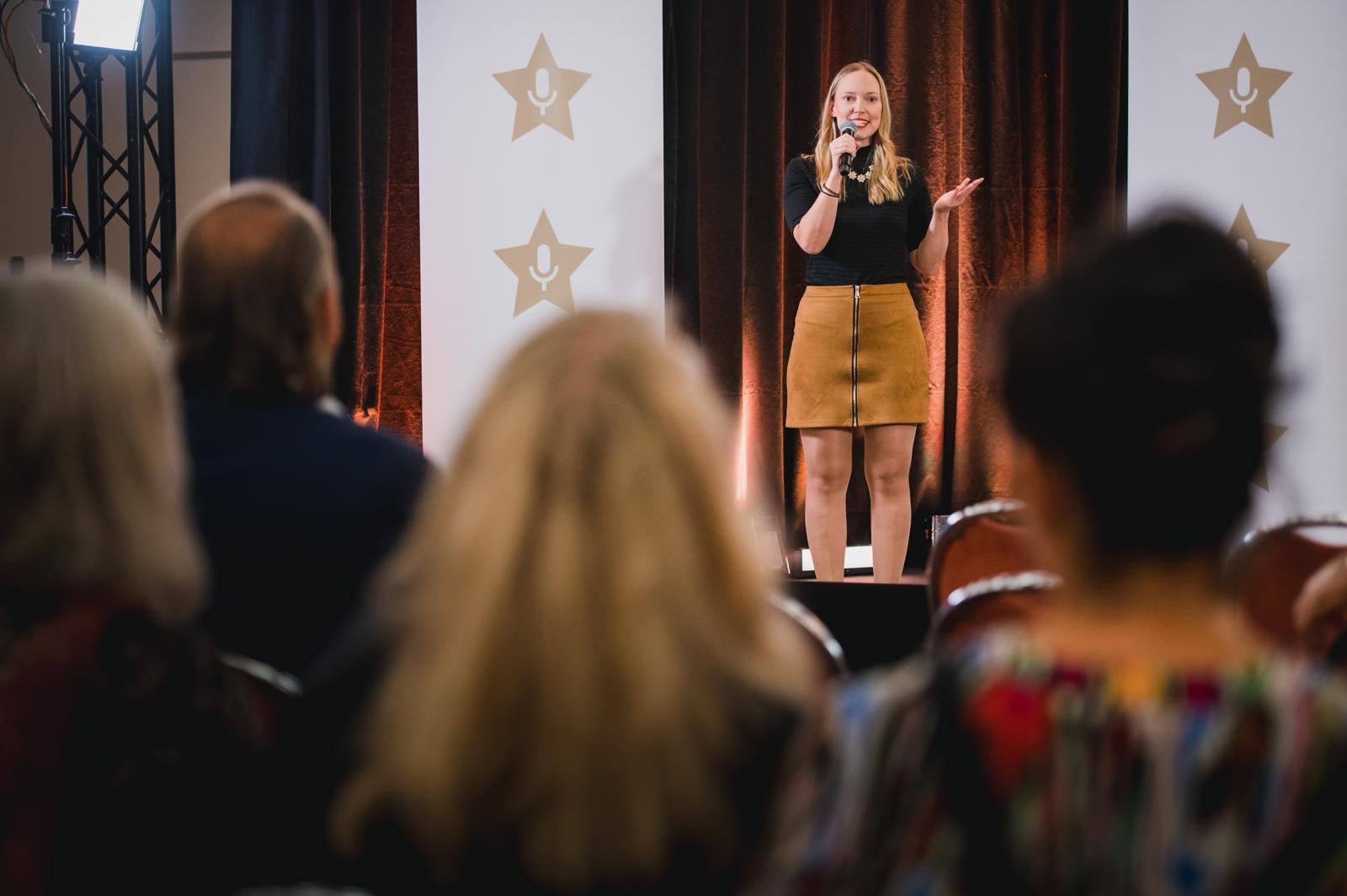 A woman speaks on stage to an audience in a conference setting.