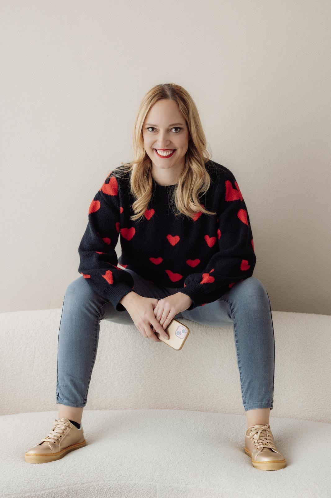 Woman sitting on a couch, wearing a heart-patterned sweater and jeans, smiling at camera