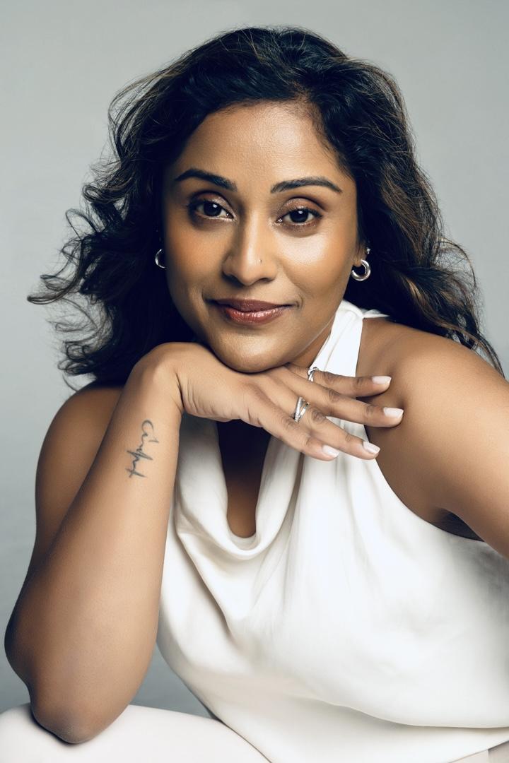 A woman with curly hair poses, resting her chin on her hand, wearing a white top