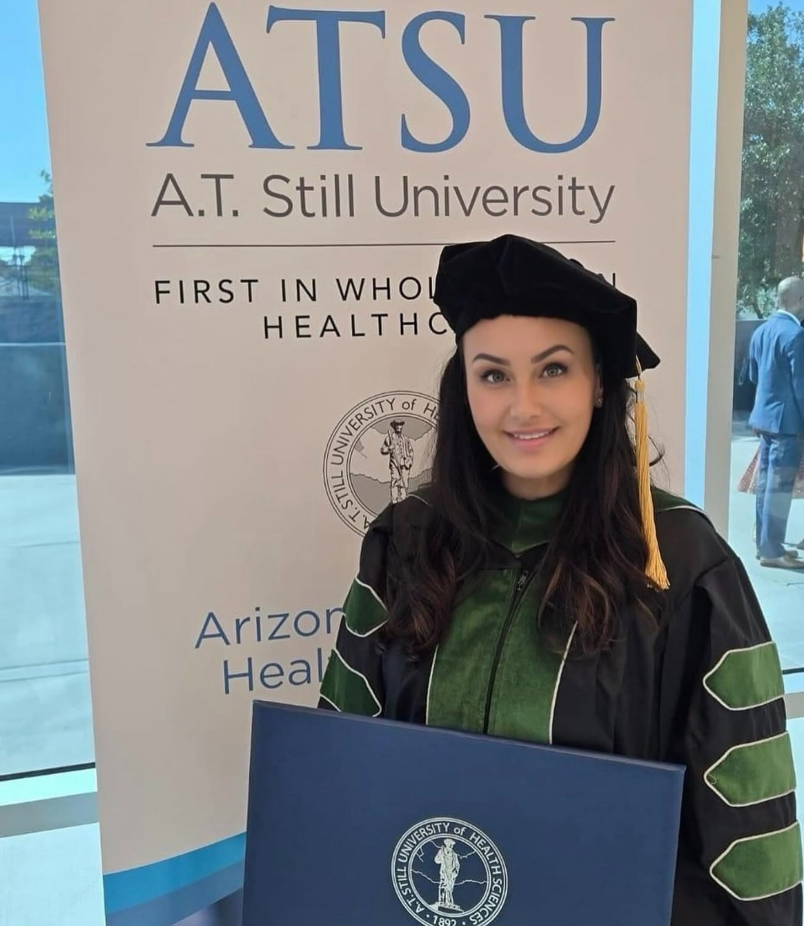 Graduate in academic regalia holding diploma with A.T. Still University banner in background