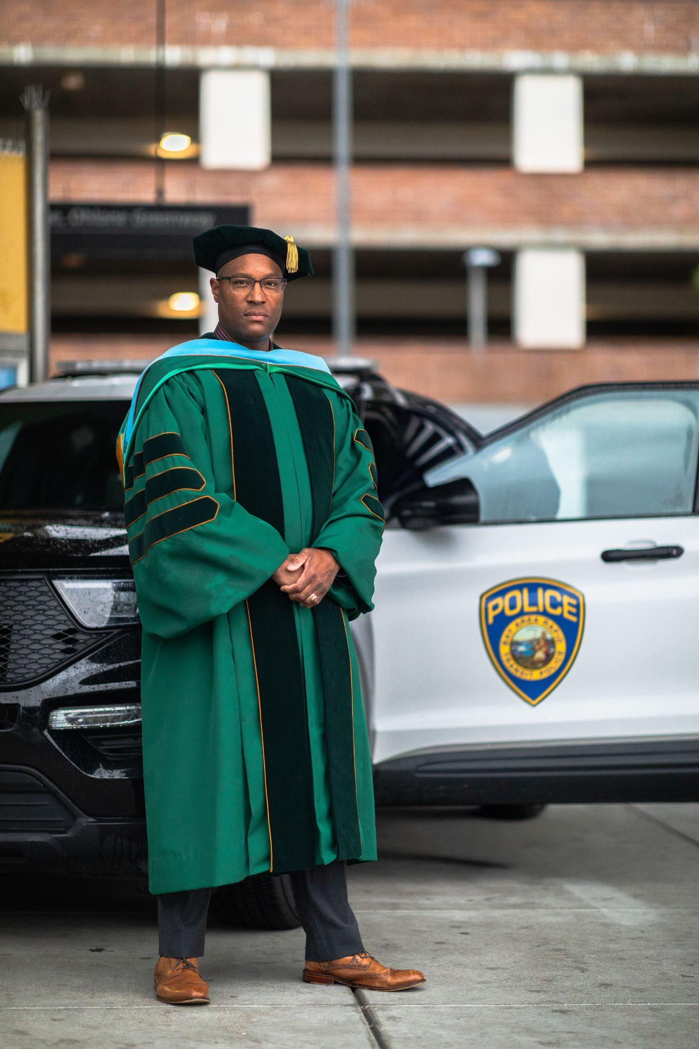 Person in academic regalia stands beside a police vehicle in a parking garage
