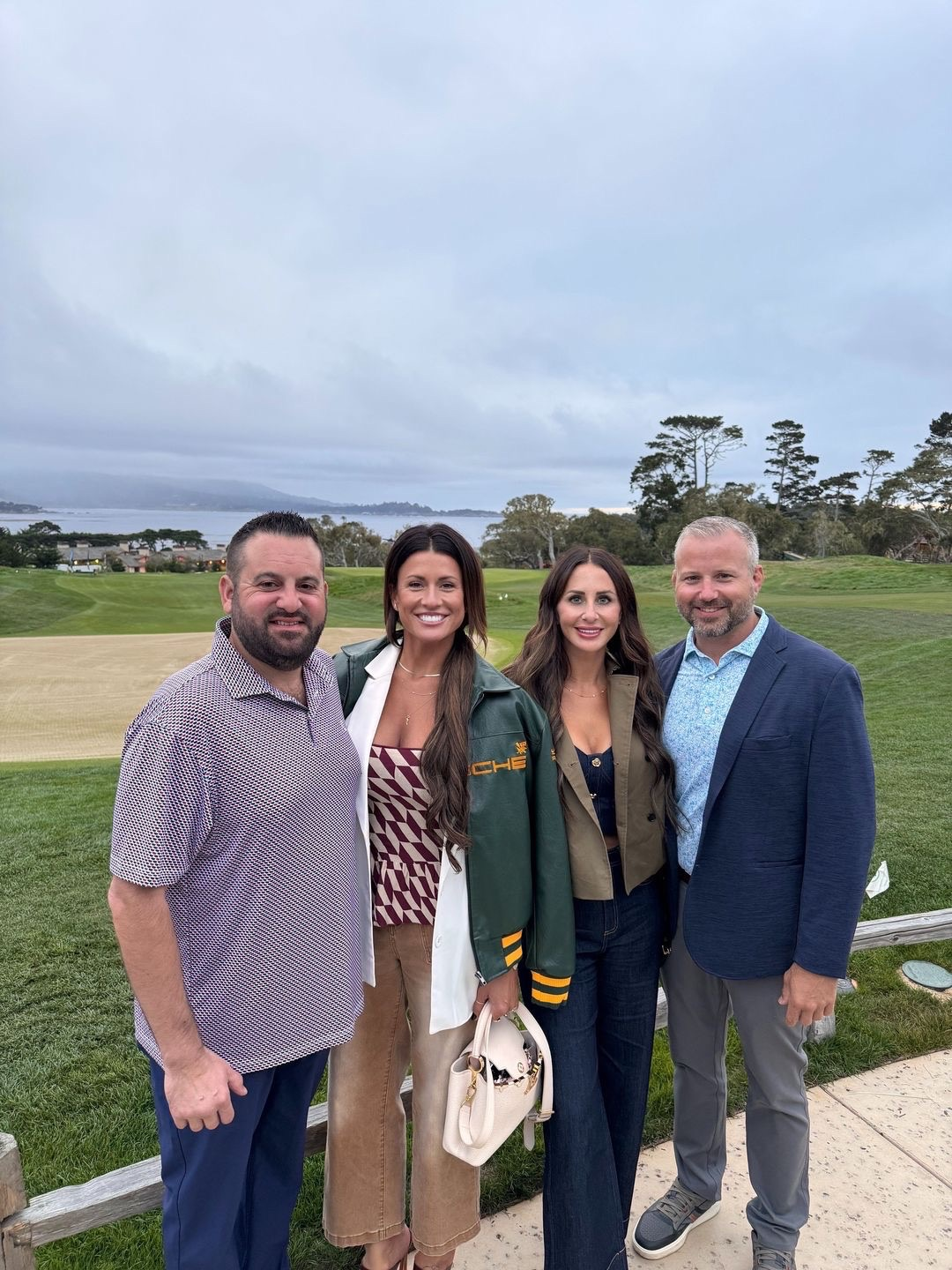 Four people pose outdoors near a golf course, dressed in casual attire.