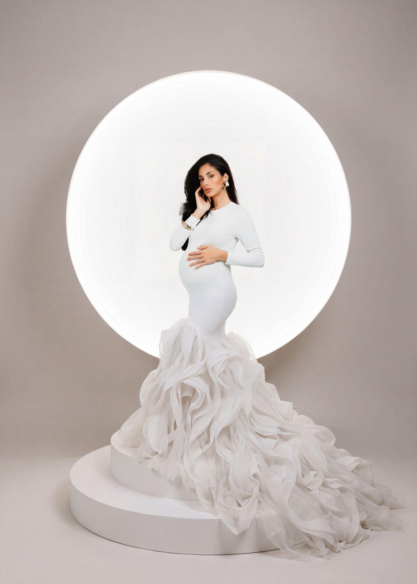 Pregnant woman in a white gown stands against a circular illuminated backdrop