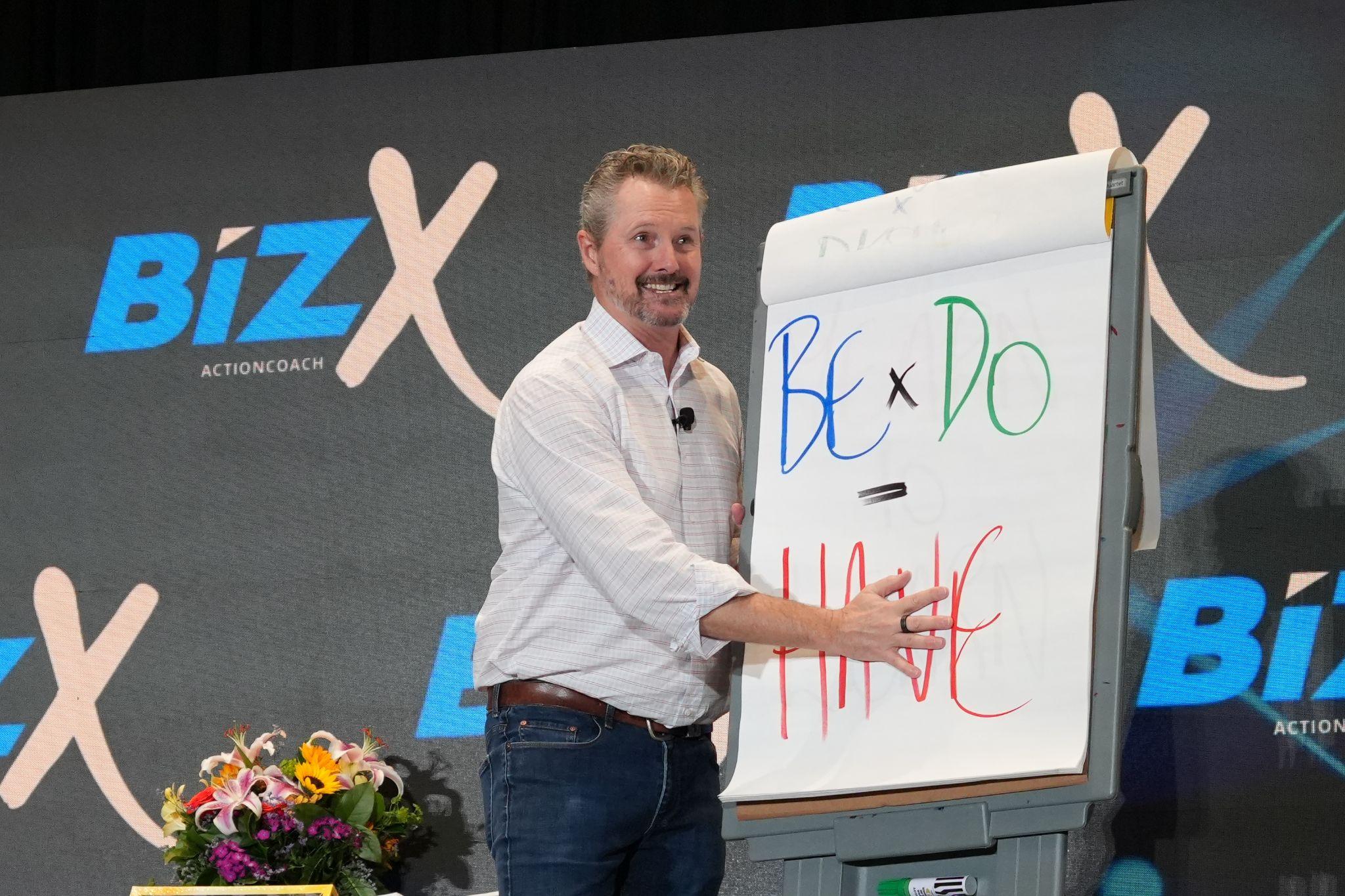 Man presenting on stage with a flip chart illustrating a formula for success