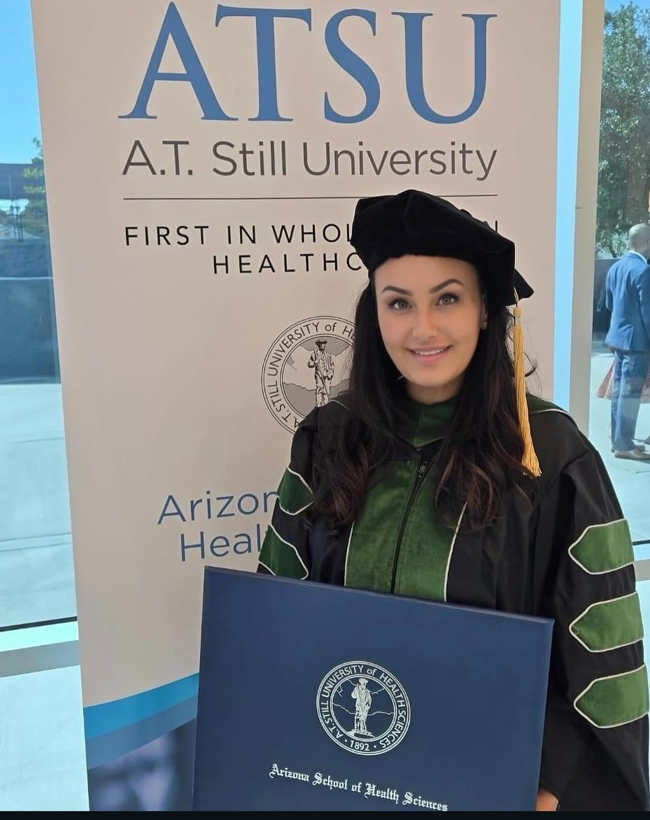 A graduate in a black cap and gown poses with a diploma at ATSU event