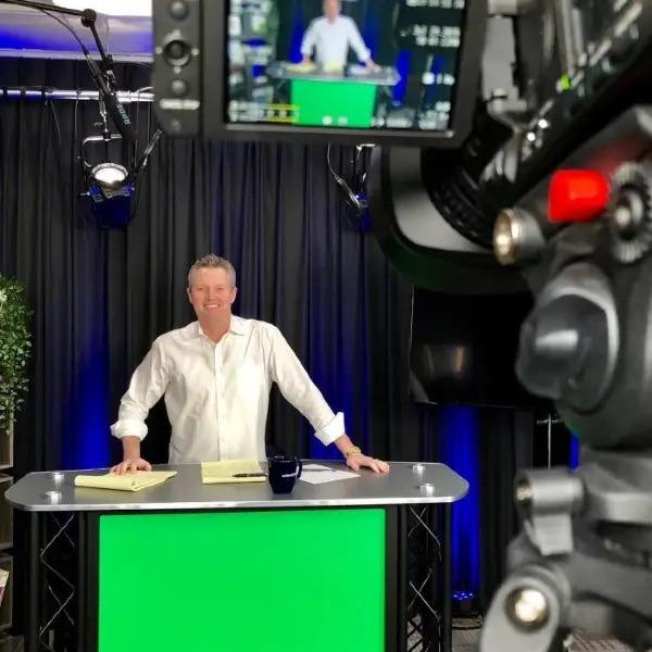 A presenter stands at a desk with a green screen background and camera setup