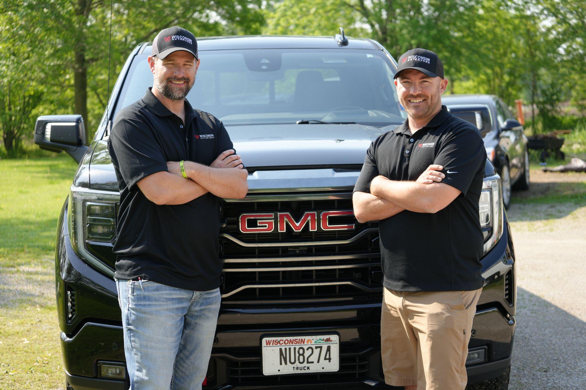 Two men in black shirts standing in front of a GMC truck in a green outdoor setting