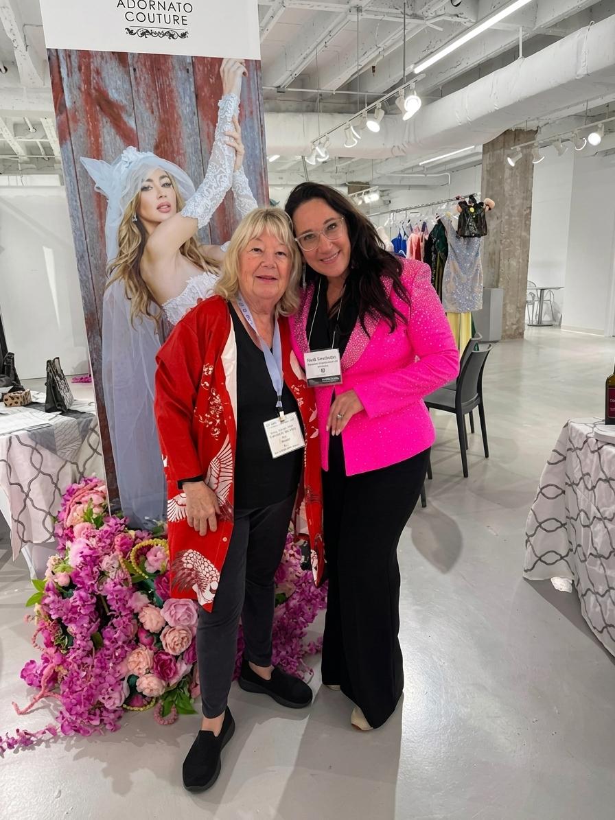 Two women pose for a photo at a fashion event with flowers and banners
