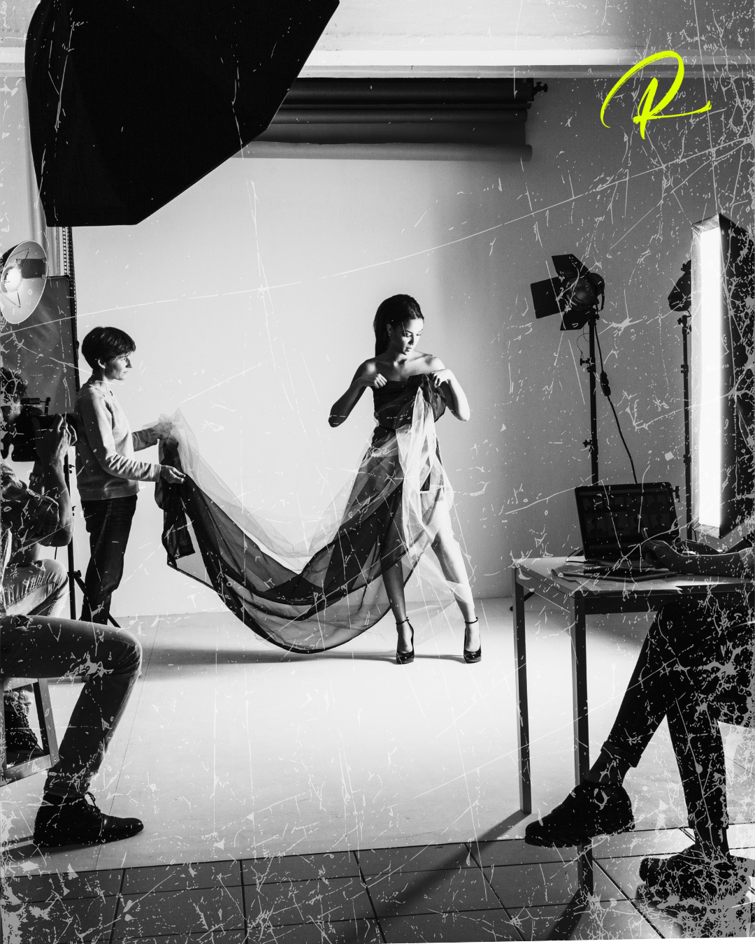 A model poses with flowing fabric while a photographer adjusts lighting in a studio.