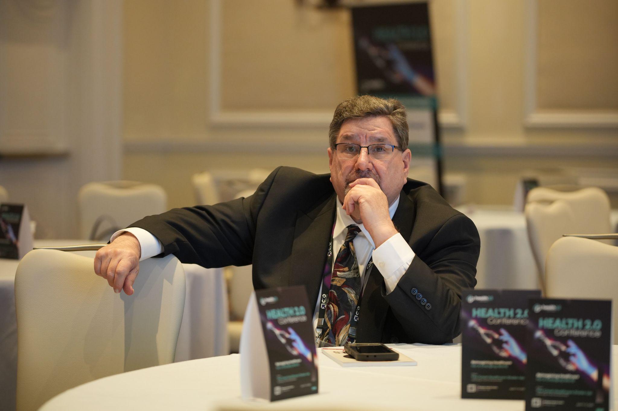 A man in a suit sits thoughtfully at a round table in a conference setting