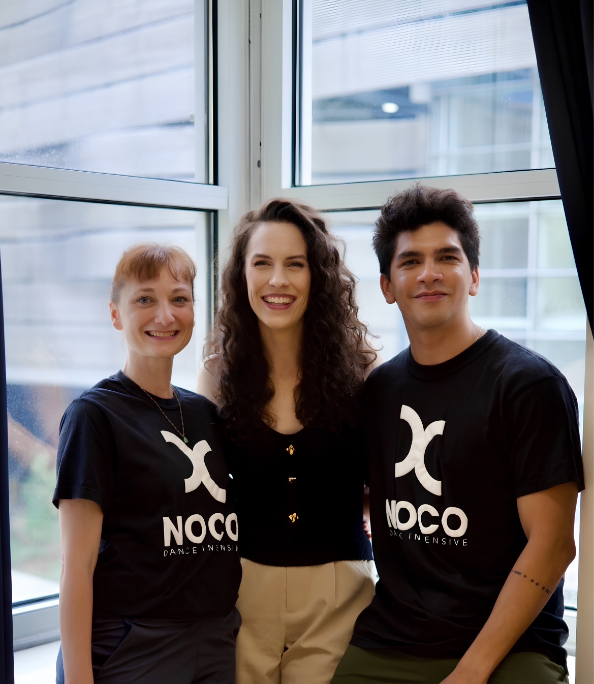 Three individuals pose together in front of a large window indoors, smiling happily