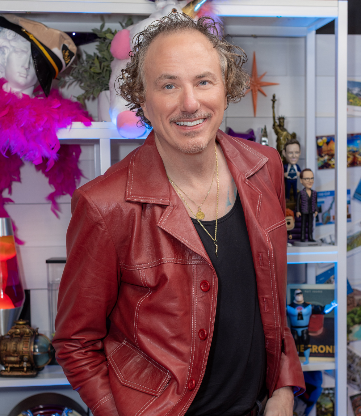 Man with curly hair in a red leather jacket smiles in a colorful setting