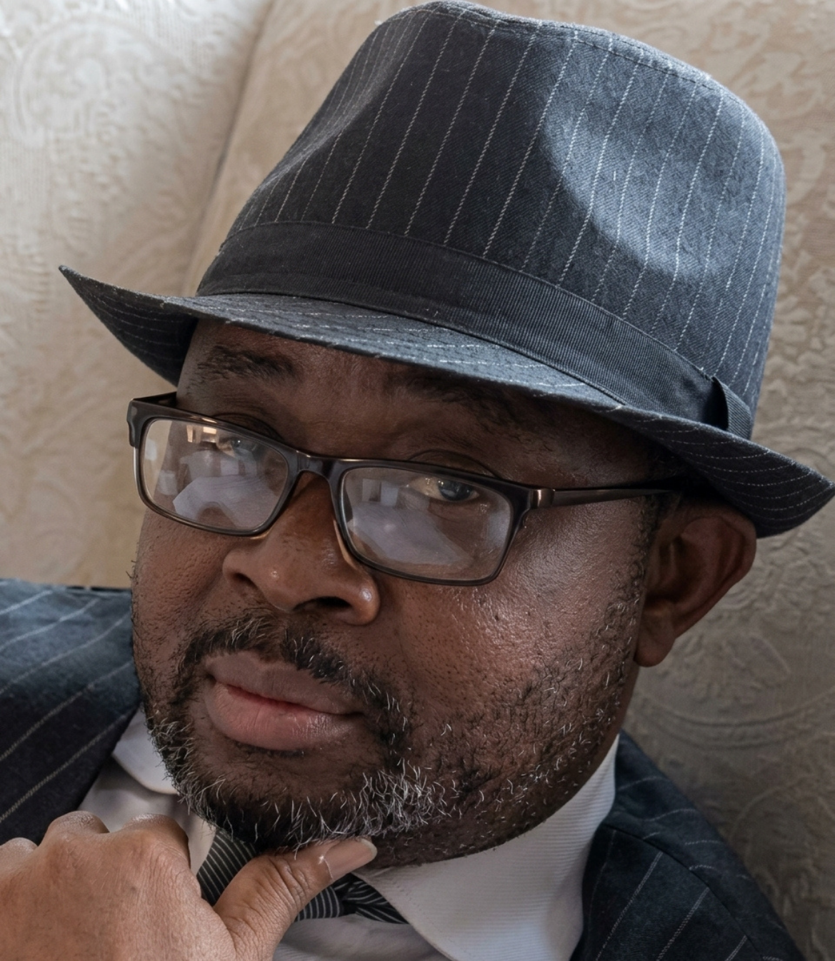 Man in a pinstripe suit and fedora hat, looking thoughtfully at the camera