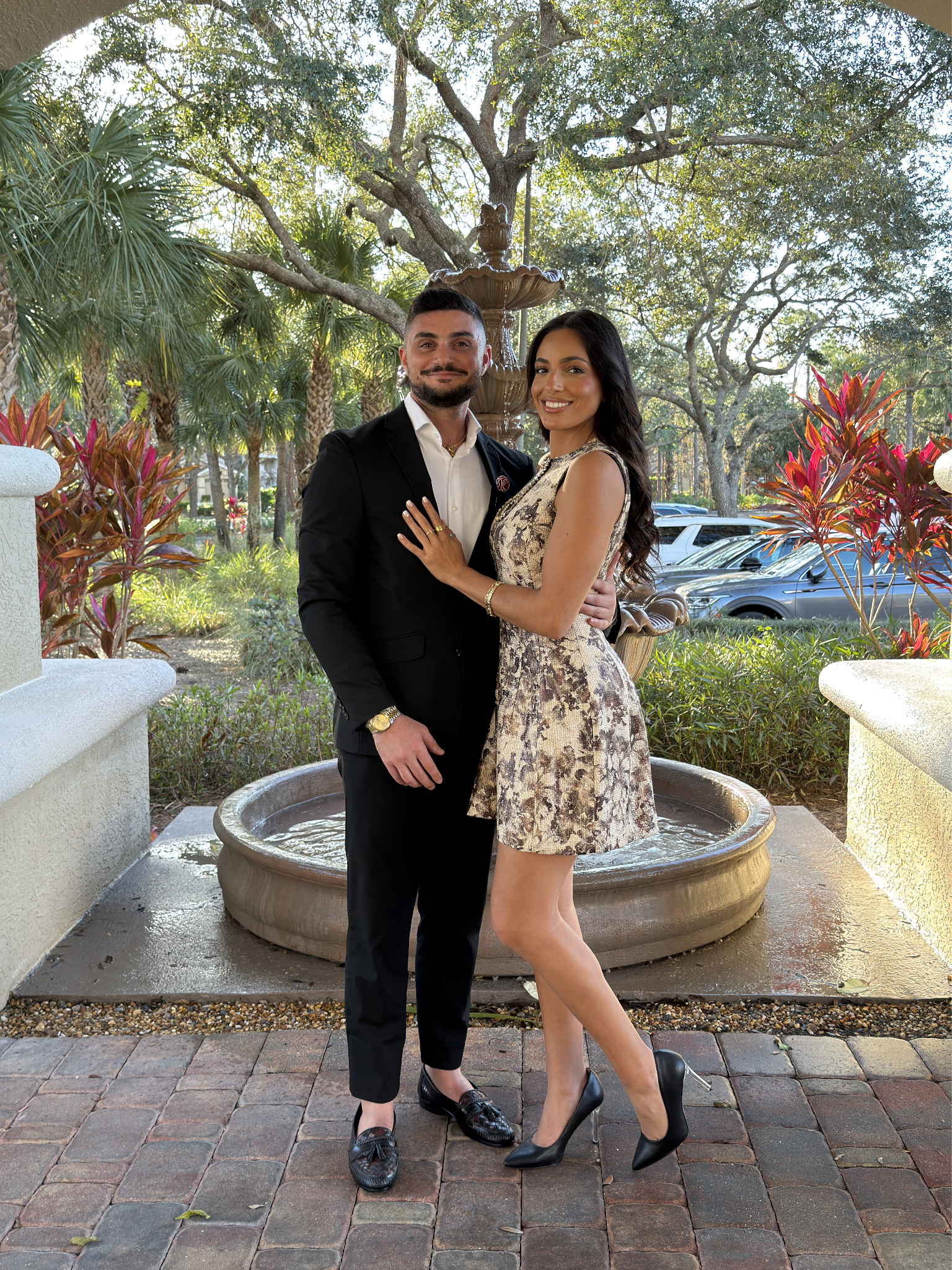 A couple poses together outside, near a fountain, dressed in formal and floral attire