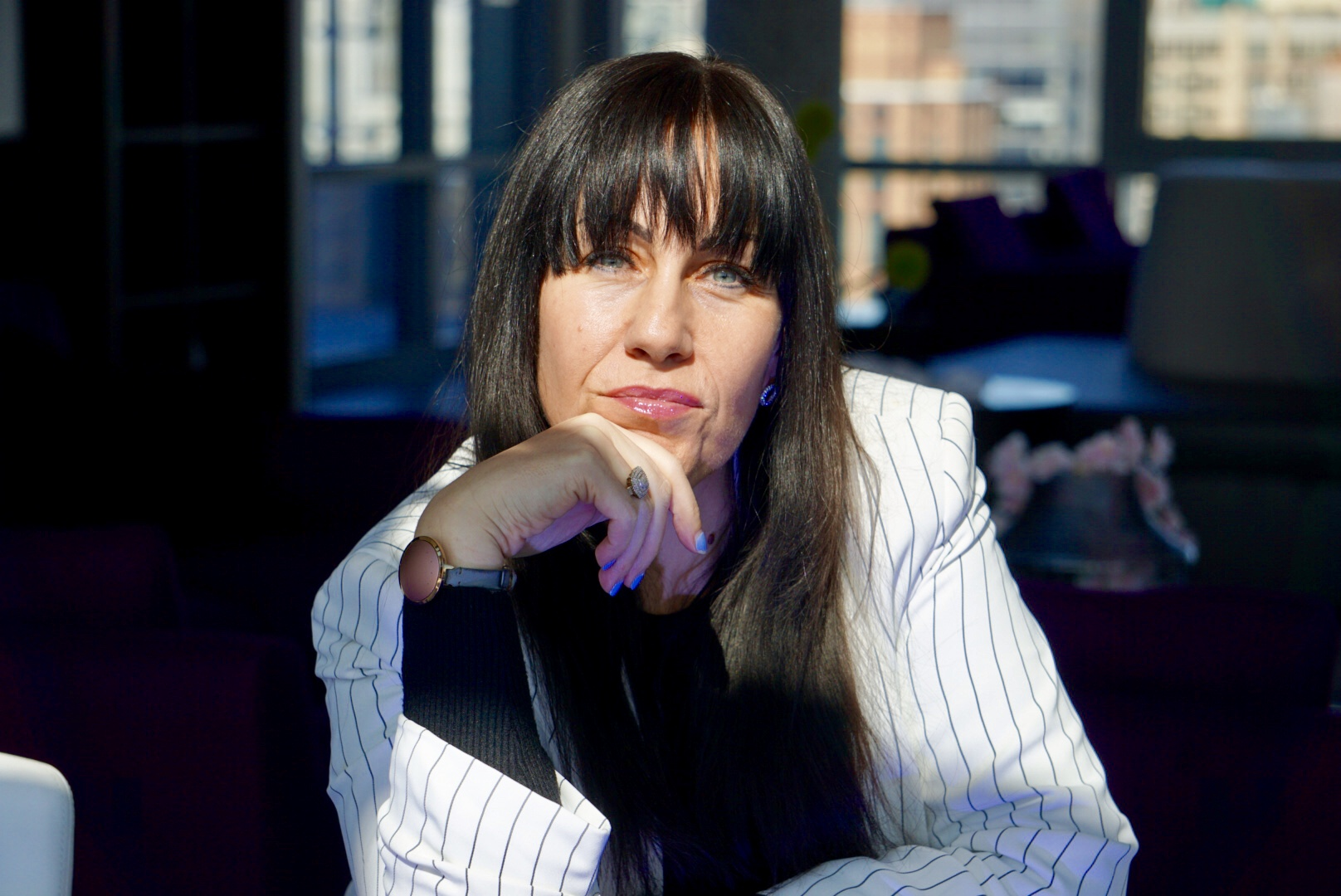 A woman with long black hair, wearing a pinstriped blazer, resting her chin on her hand.