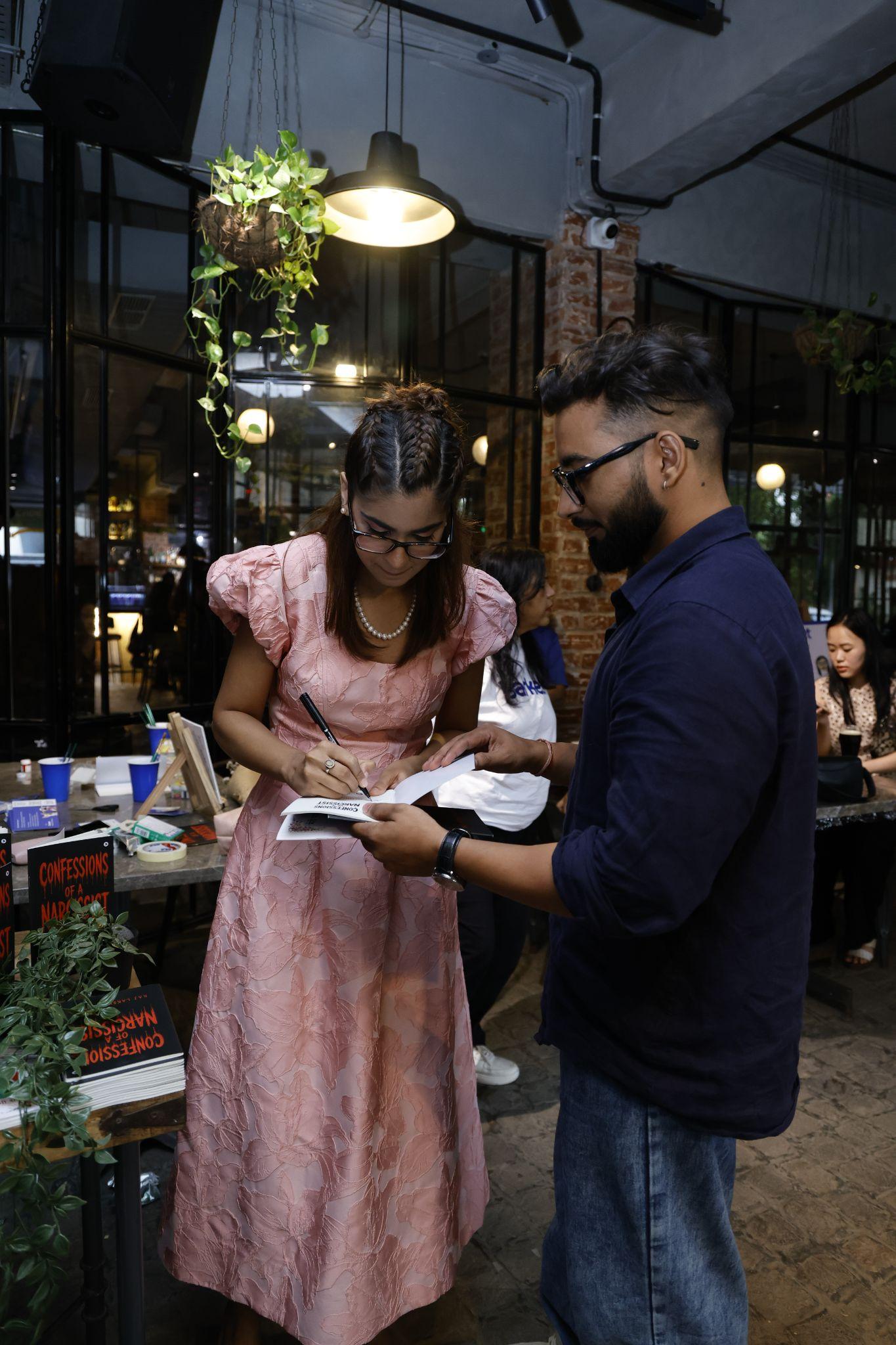 A woman in a pink dress signs an item for a man in glasses.