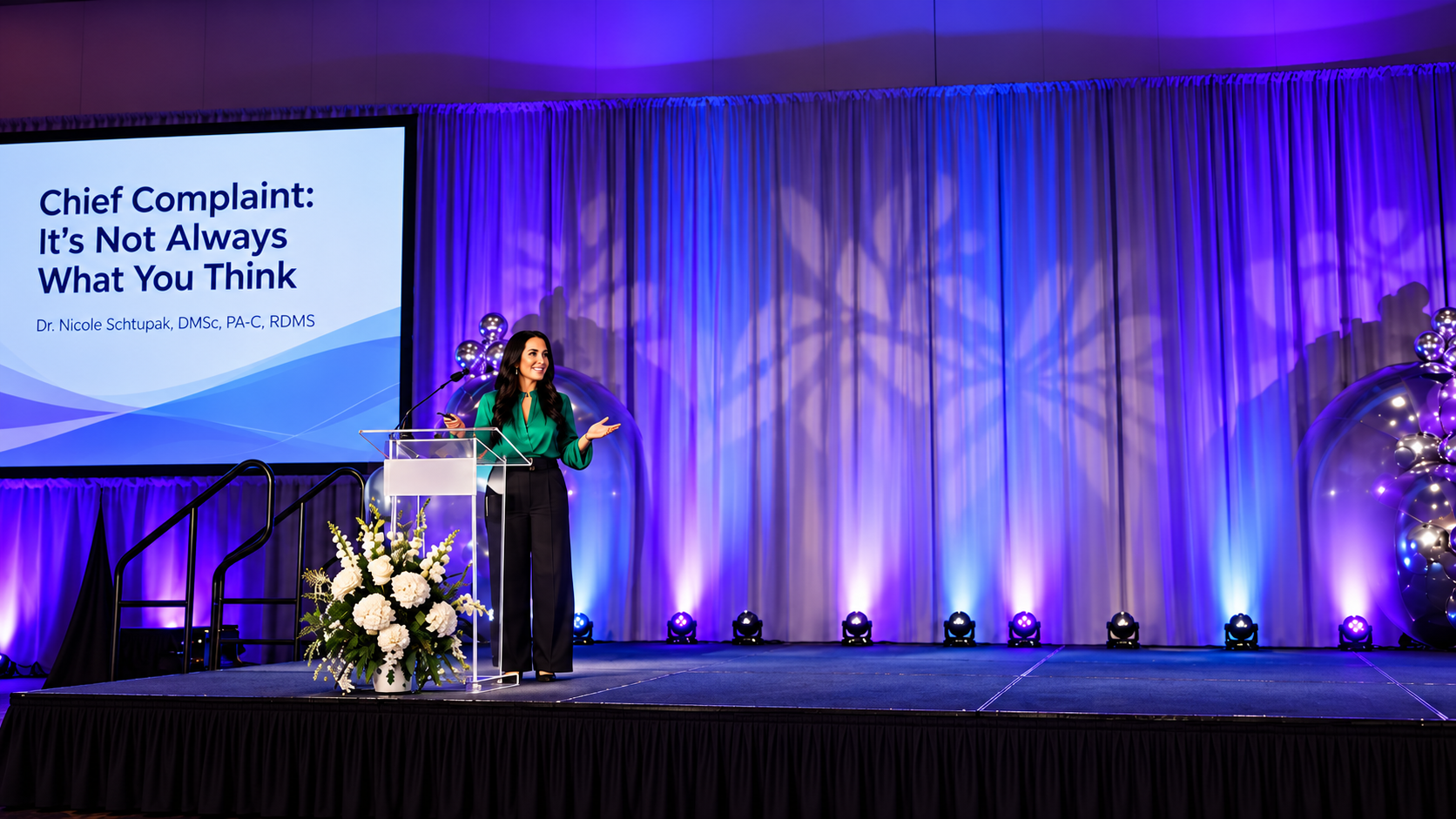 A speaker presents on stage at a conference with a colorful backdrop and floral arrangements