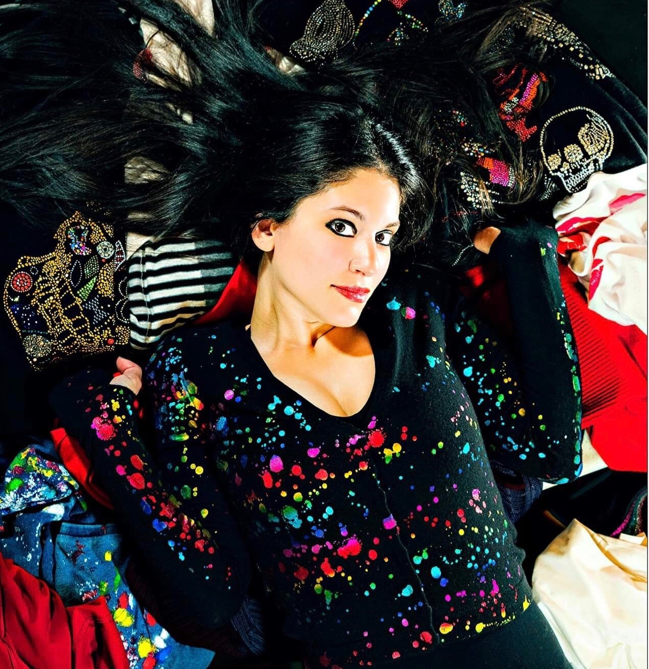 A woman with long hair lies on colorful clothes, wearing a speckled black cardigan